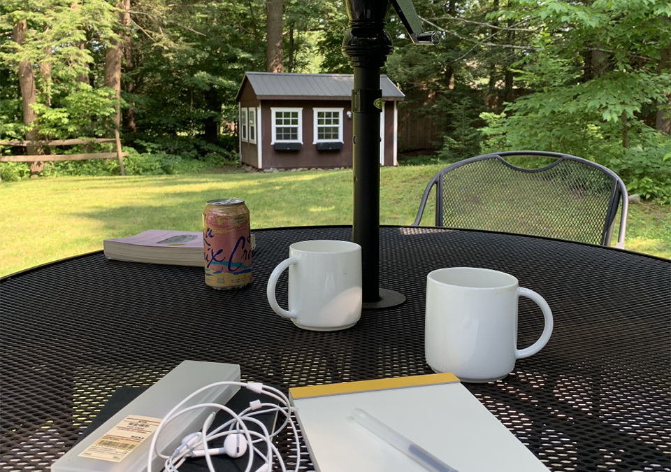 A picture of a table outdoors overlooking a yard. White mugs, a book, and a notebook sit on the table.